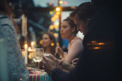 Cropped image of young man using mobile phone while sitting amidst friends at dining table during dinner party