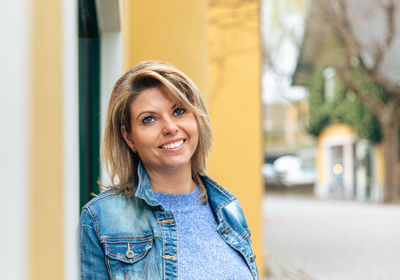Joyful woman standing by a vibrant yellow wall in a blue sweater and denim jacket