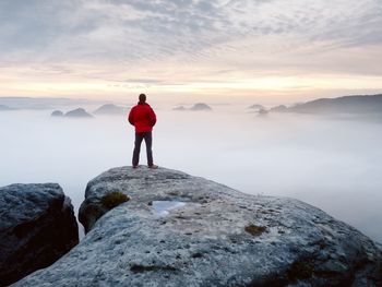 Man standing on rock looking at shore against sky during sunset