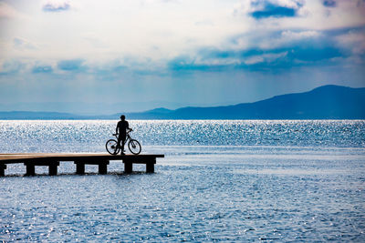 Silhouette man standing with bicycle on pier over sea against cloudy sky