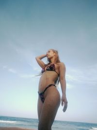 Low angle view of young woman wearing bikini while standing at beach against sky