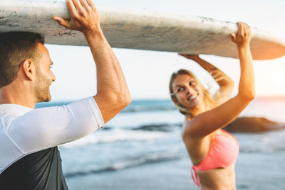 Cheerful man and woman with surfboard standing at beach against sky