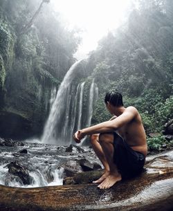 Side view of shirtless man sitting against waterfall in forest