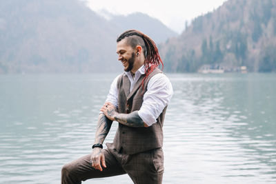 A tall brutal man hipster groom in a wedding suit stands by the misty mountains and lake in nature