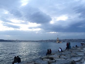 People on rocks by sea against cloudy sky