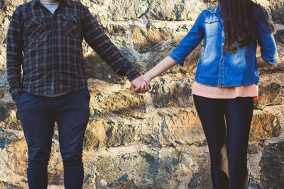 Midsection of couple holding hands while standing against stone wall