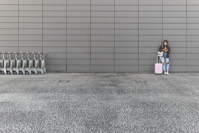 Young woman wearing mask with luggage standing against wall outside railroad station