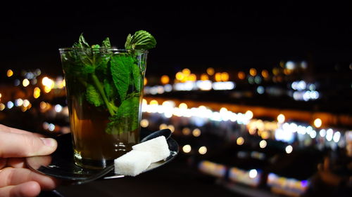 Cropped hand holding drink against illuminated city at night