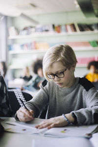 Schoolboy writing in book while sitting at desk in classroom