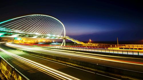 Light trails on street by bridge against sky at night