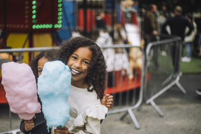 Portrait of happy girl enjoying with sister holding cotton candies at amusement park