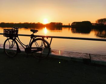 Silhouette bicycle by lake against sky during sunset