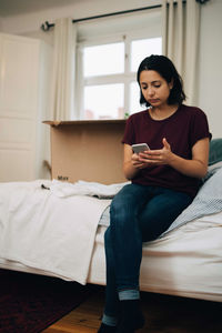 Woman using mobile phone while sitting by box on bed