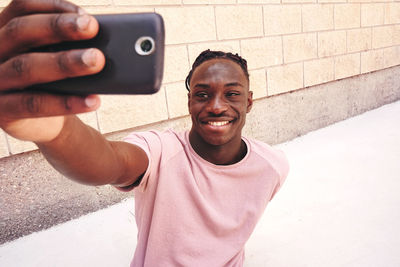 Smiling teenage boy taking selfie with smart phone while standing against wall