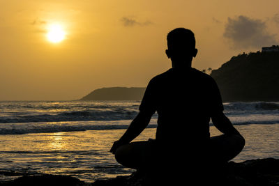 Silhouette man meditating on beach against sky during sunset