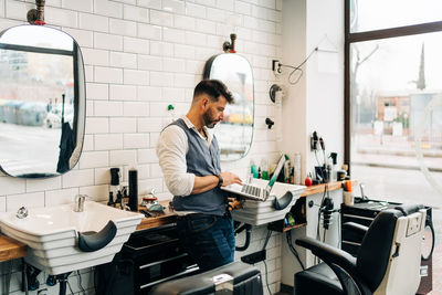 Side view of masculine male hairdresser browsing internet on netbook in modern beauty salon against window