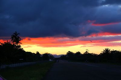 Road by silhouette trees against dramatic sky