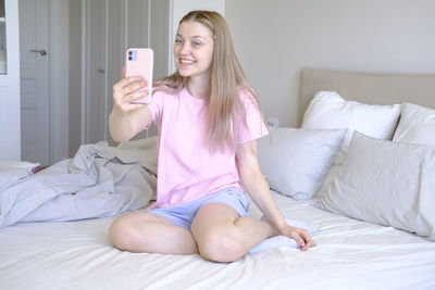 Cute girl 15-18 years old sitting on the bed makes a selfie.
