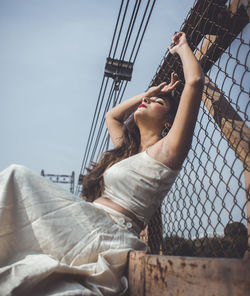 Low angle view of young woman sitting by fence against clear sky