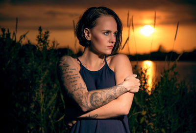 Young woman looking away while standing against plants during sunset