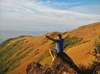 Man with arms outstretched sitting on rock against sky