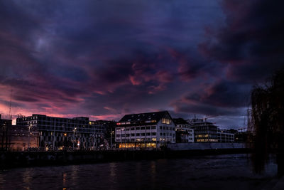 Illuminated buildings by river against sky at sunset