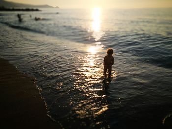 Silhouette baby girl standing in sea on shore at beach during sunset