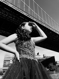 Low angle view of girl shielding eyes while standing against bridge during sunny day