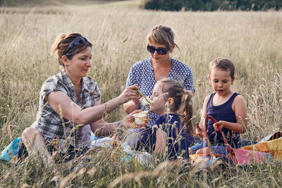 Woman feeding daughter while sitting with friend on grassy field