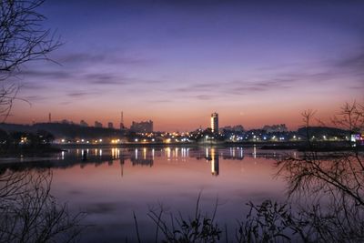 Illuminated city by lake against sky at sunrise