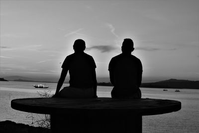 Rear view of silhouette men sitting by sea against sky