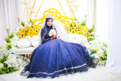 Portrait of beautiful bride holding flower bouquet while sitting on sofa 