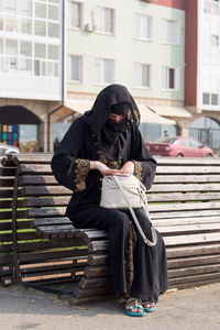 A muslim woman in national clothes sitting bench is looking for something her women's handbag.