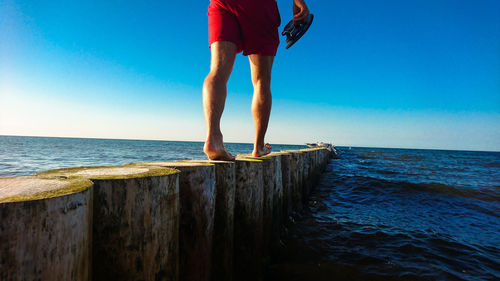Low section of man walking on wooden posts in sea against blue sky