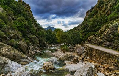 Stream flowing through rocks by river against sky