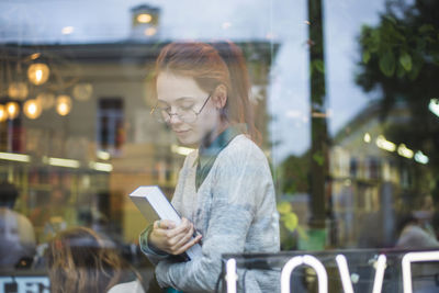 Through window side view of young stylish female in eyeglasses sitting in cozy bookshop with volume
