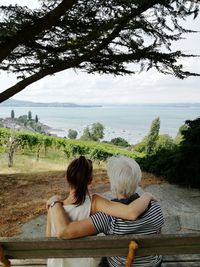 Rear view of grandmother and granddaughter sitting on bench against sea