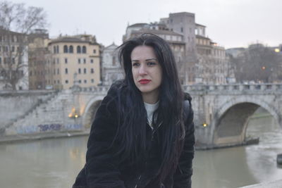 Young woman looking away while sitting against river in city