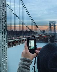 Midsection of person photographing with suspension bridge against sky