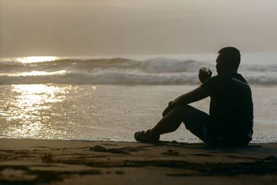 Man sitting on shore at beach against sky during sunset