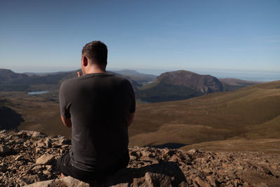 Rear view of man sitting on mountain against sky