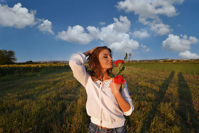 Young woman holding flower standing on field against sky