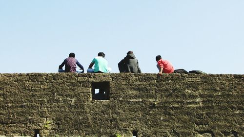 Rear view of friends sitting on surrounding wall against sky