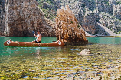 Side view of shirtless man sitting on rowboat in sea against rock formations