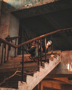 Low angle view of man wearing mask sitting on stairs in abandoned building