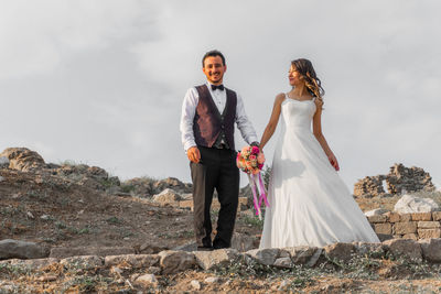 Portrait of smiling bride with groom holding bouquet while standing on footpath against sky