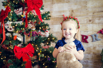 Cute girl holding teddy bear standing by decorated christmas tree against wall at home