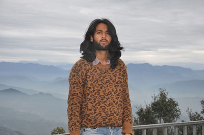 A good looking indian young man with long hair and beard looking at camera while standing in hill