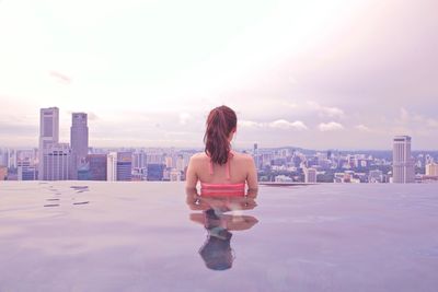 Rear view of woman standing in infinity pool against buildings in city