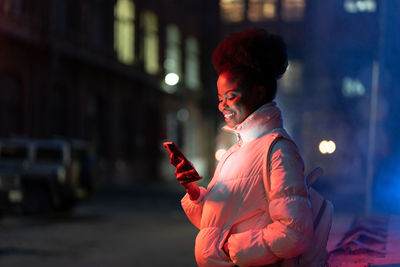 Happy african woman looking at screen of mobile phone while standing outdoors on cold winter night